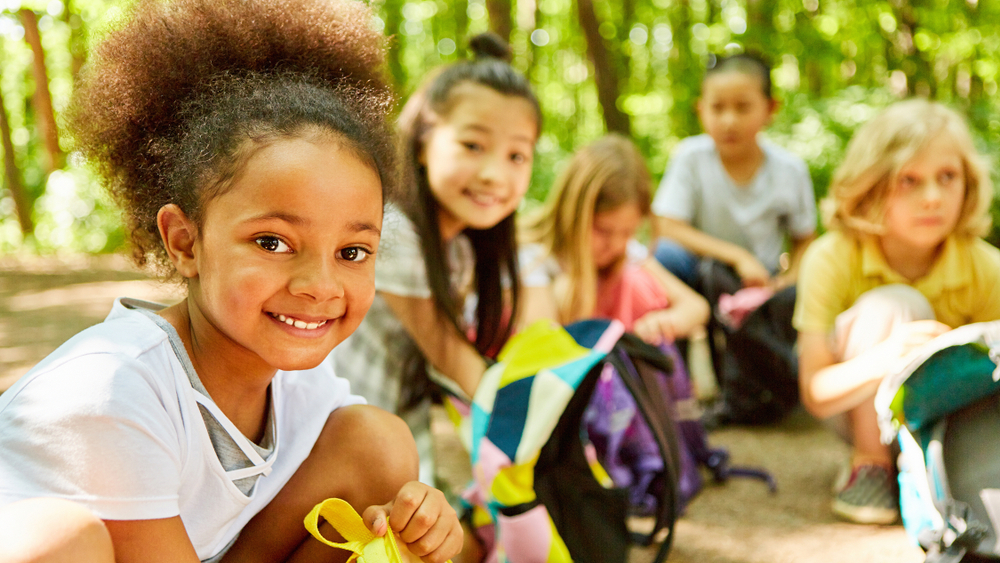 A group of five children sitting on a forest trail, with one smiling girl in focus.